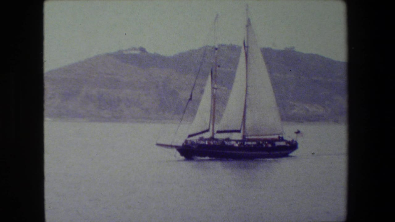 The sailboat remains centered in the frame, its white sails standing against muted water and a soft, distant hillside.