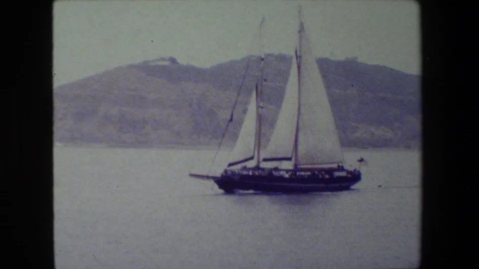 The sailboat remains centered in the frame, its white sails standing against muted water and a soft, distant hillside.