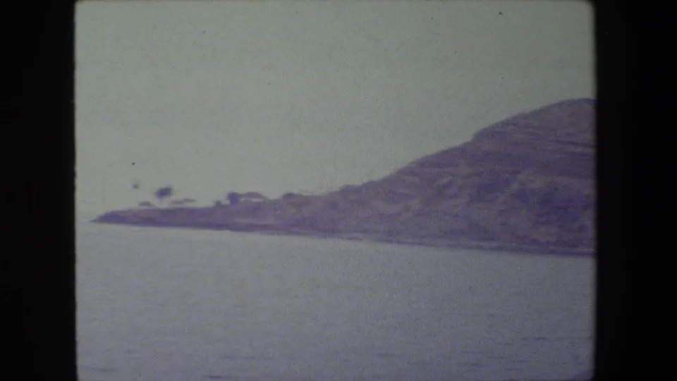 A soft 8mm frame shows calm water in the foreground and a dry sloping hillside across the shoreline, with no people or structures visible.
