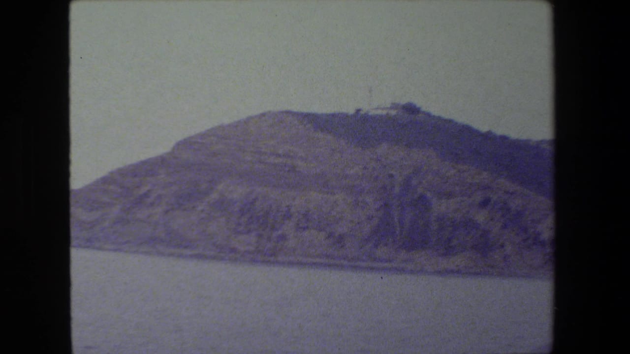 The pan frames a broad, rugged hillside above still water, with a muted purple-blue film cast and no identifiable landmark visible.