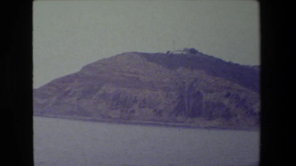 The pan frames a broad, rugged hillside above still water, with a muted purple-blue film cast and no identifiable landmark visible.