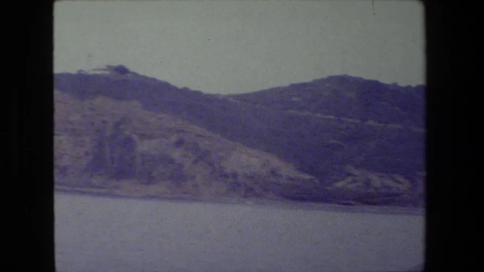 A closer 8mm view shows textured rocky slopes dropping toward the waterline, framed by the camera pan and softened by home-movie grain.