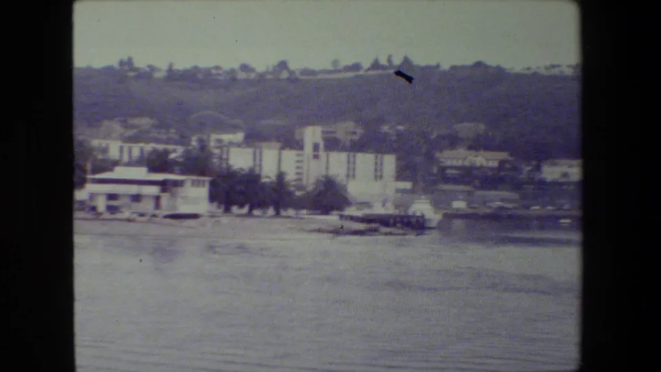 A soft handheld 8mm frame shows pale waterfront structures, trees, and a low hillside beyond the water, with no visible people or readable markings.