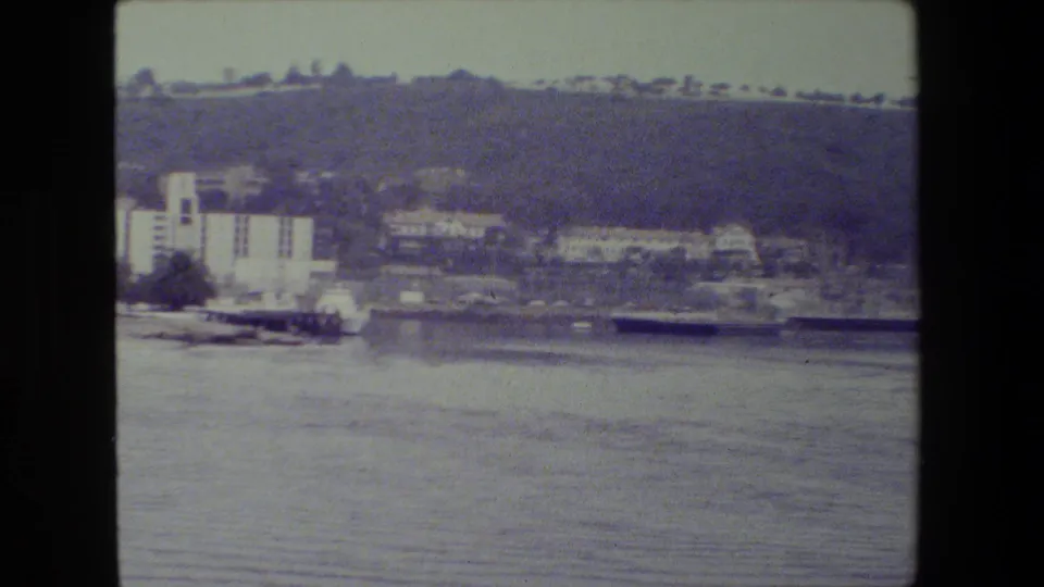 Across the harbor, distant buildings, a dock or pier edge, trees, and a hillside are visible in a muted 8mm home-movie frame.