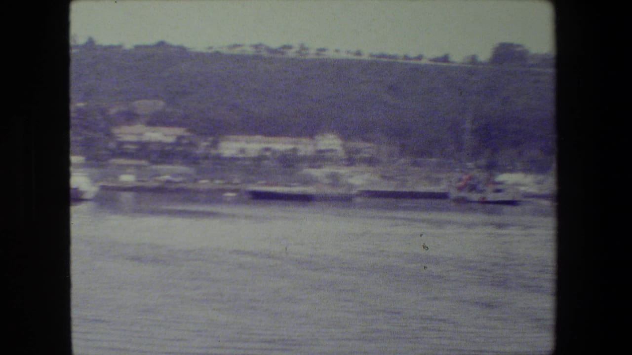 Soft 8mm frame of a naval vessel seen from across calm water, with shoreline buildings and a tree-covered hillside in the background.