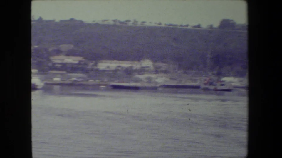 Soft 8mm frame of a naval vessel seen from across calm water, with shoreline buildings and a tree-covered hillside in the background.
