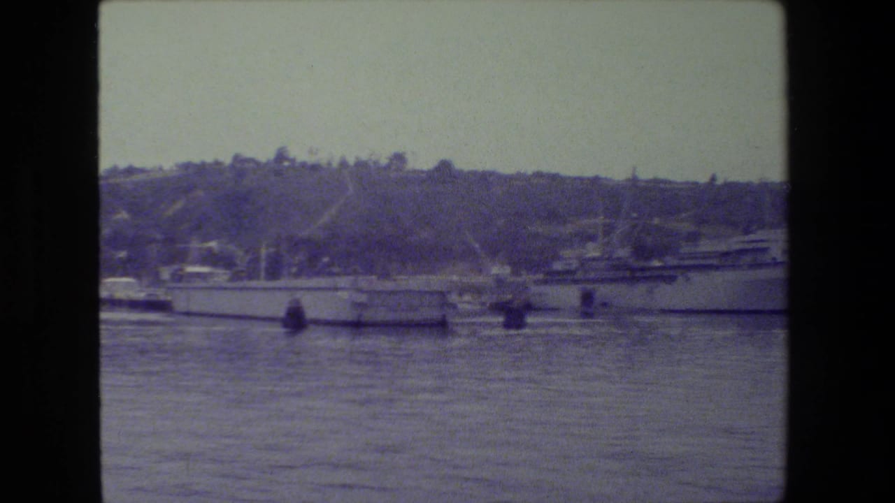 A large gray naval ship appears moored in a harbor near smaller boats, with a wooded hillside and waterfront structures behind it.