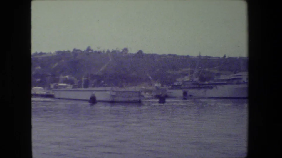 A large gray naval ship appears moored in a harbor near smaller boats, with a wooded hillside and waterfront structures behind it.
