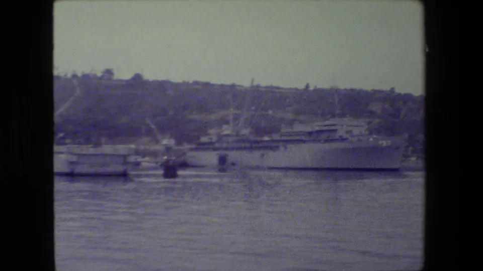 The camera frames more of the naval vessel's side, including the visible hull number 37, with dock structures and hillside beyond.