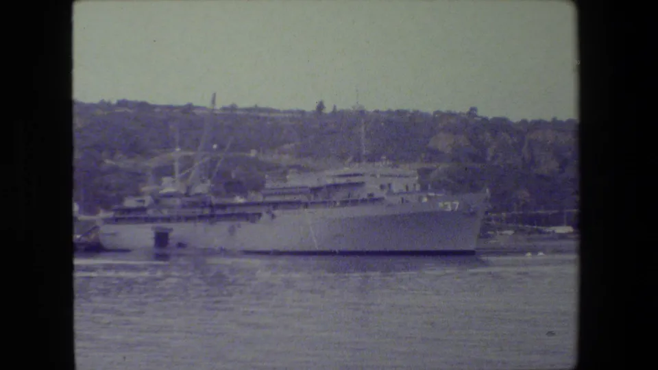 Broadside 8mm view of a gray naval ship with hull number 37, moored in calm harbor water below a tree-covered shoreline.