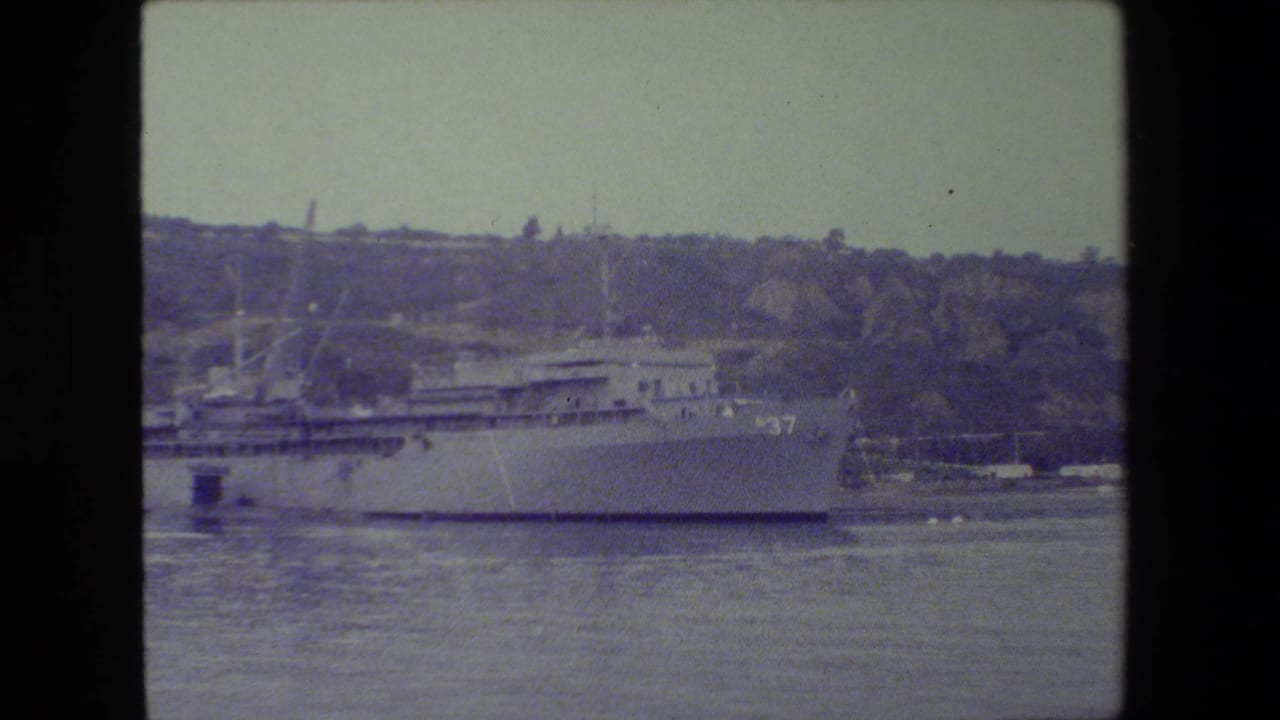 A large gray naval vessel appears docked across calm harbor water, with a wooded hillside and shoreline structures behind it in archival 8mm footage.