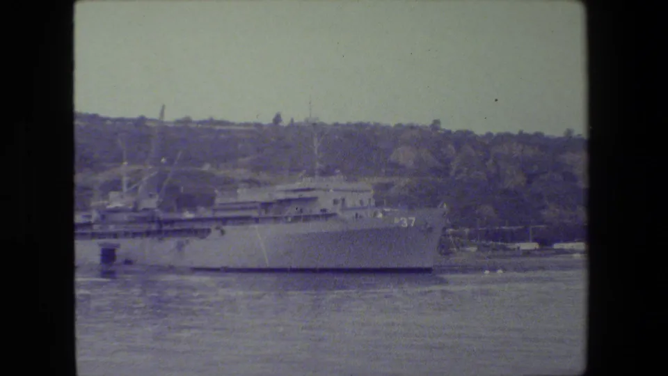 A large gray naval vessel appears docked across calm harbor water, with a wooded hillside and shoreline structures behind it in archival 8mm footage.