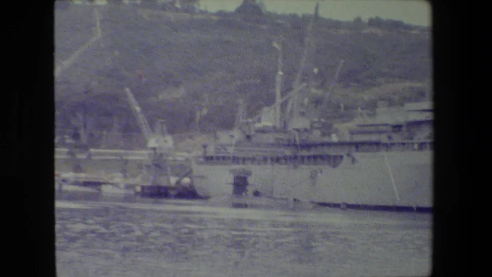 The frame moves closer to the pier area, showing cranes, dock structures, and part of a large ship along the waterfront in soft, tinted 8mm footage.