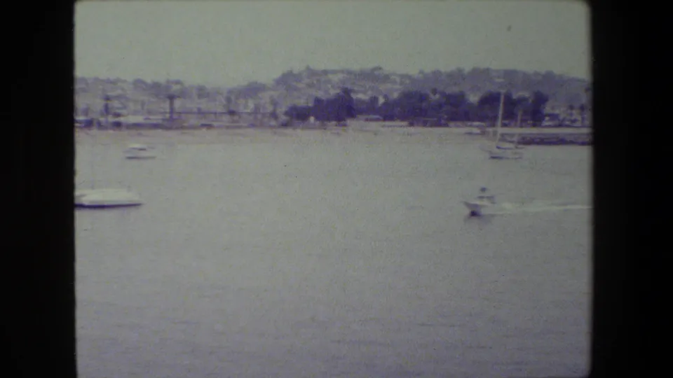 Several small boats move or sit on open harbor water, with a distant shoreline, low buildings, and hillside visible through faded 8mm color.