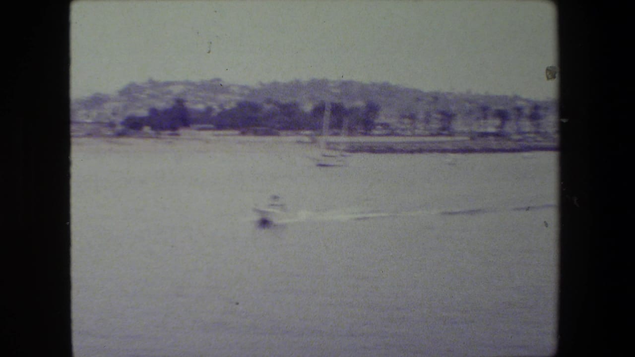 A small motorboat moves across calm harbor water in archival 8mm footage cataloged near San Diego Naval Base, with sailboats and a hazy shoreline in the background.