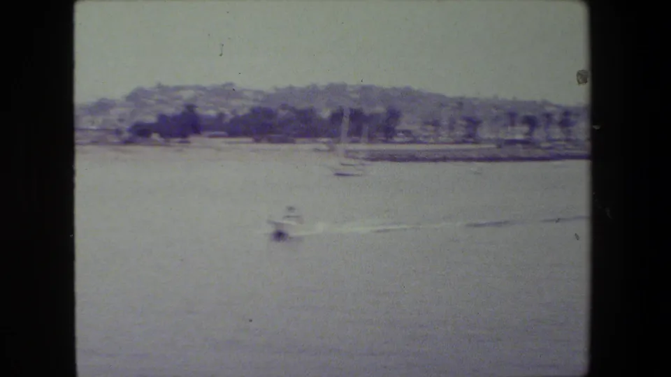 A small motorboat moves across calm harbor water in archival 8mm footage cataloged near San Diego Naval Base, with sailboats and a hazy shoreline in the background.