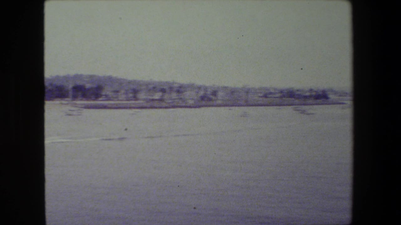 Calm harbor view with a faint motorboat wake across the water, a low shoreline, and small boats visible under a hazy sky.