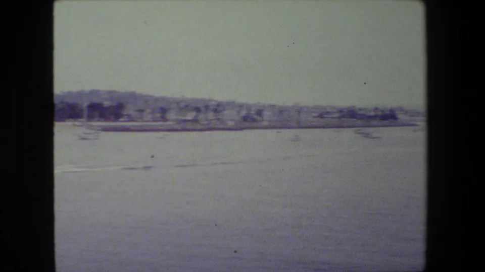 Calm harbor view with a faint motorboat wake across the water, a low shoreline, and small boats visible under a hazy sky.