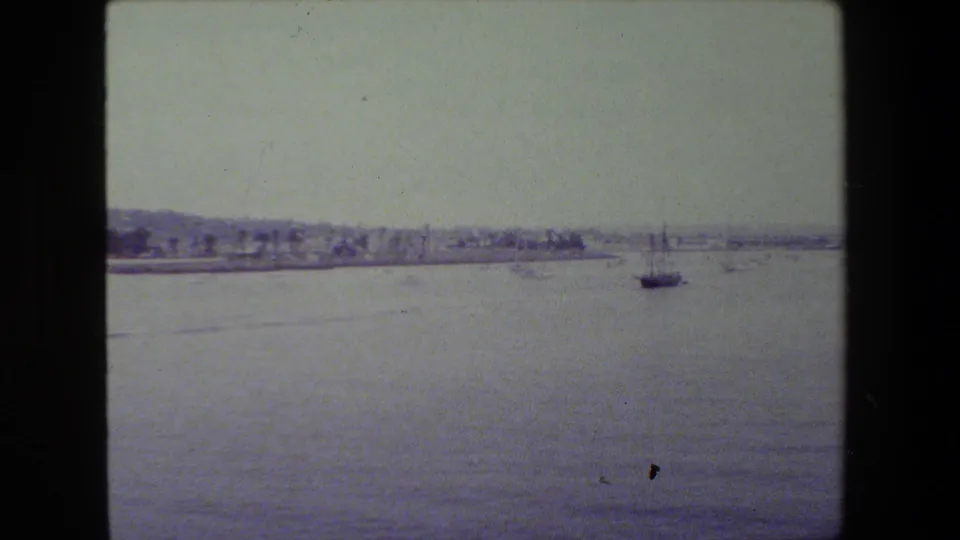 Wide 8mm harbor frame showing open water, a low waterfront shoreline, and a dark sailboat positioned near the right side of the frame.