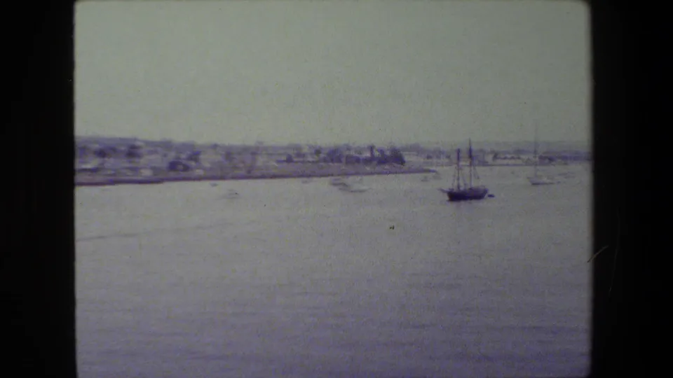 A larger sailboat sits on calm harbor water in soft 8mm footage, with distant shoreline buildings and a hazy coastal sky behind it.