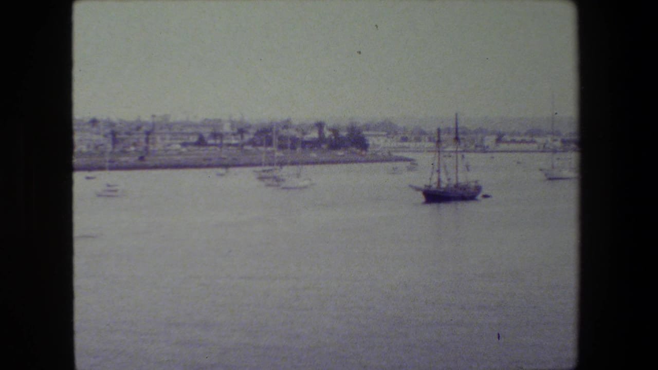 A wide 8mm harbor view shows calm water, a small sailboat near center, distant waterfront buildings, and dark film-scan borders.