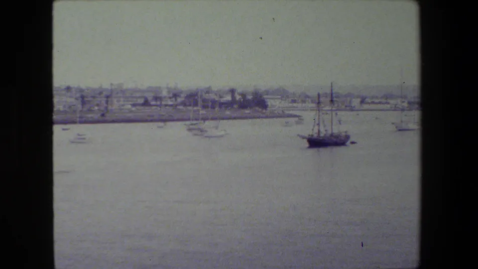 A wide 8mm harbor view shows calm water, a small sailboat near center, distant waterfront buildings, and dark film-scan borders.
