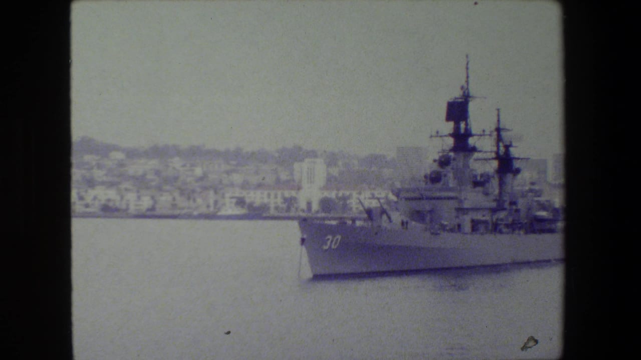Large naval warship marked 30 moving through harbor water with city buildings behind it.