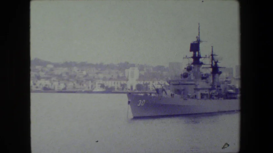 A large gray naval warship bearing the number 30 passes through calm harbor water, with a hazy urban shoreline in the background.