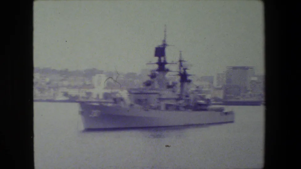 The warship appears in a softer side view as it moves across the harbor, with masts, deck structures, water, and shoreline buildings visible through faded 8mm color.