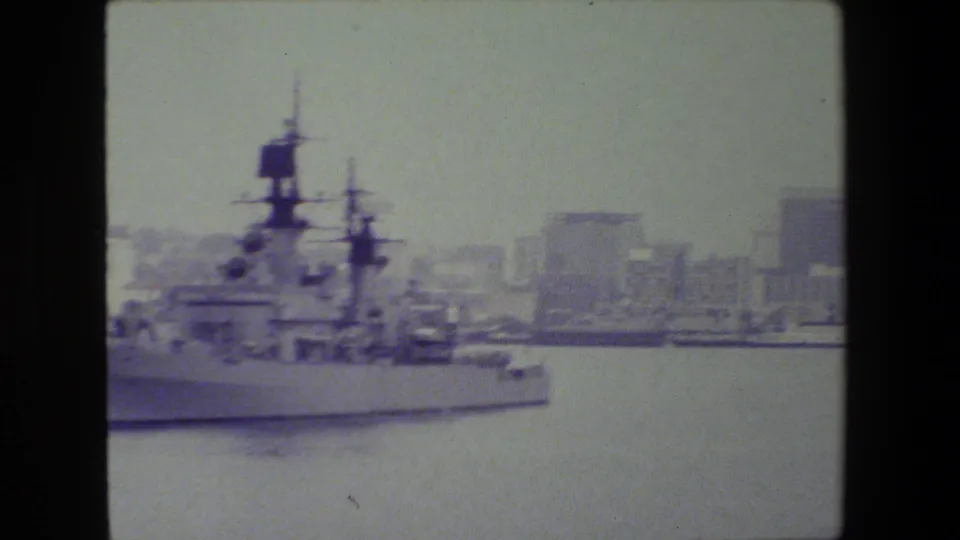 A large naval ship continues through the harbor in a soft, faded 8mm frame, with urban buildings and waterfront structures behind it.