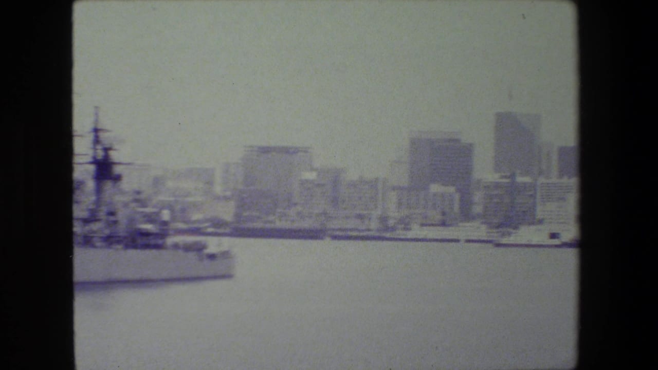 A large vessel sits along the waterfront at left while distant high-rise buildings appear across the water in faded 8mm footage reported as San Diego naval base material.