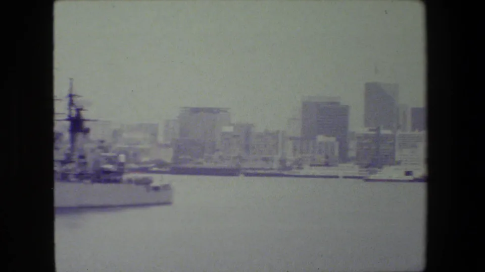A large vessel sits along the waterfront at left while distant high-rise buildings appear across the water in faded 8mm footage reported as San Diego naval base material.