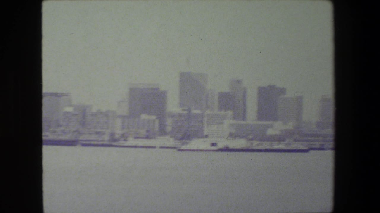 The camera frames open water and a hazy line of high-rise buildings along the waterfront, with the vessel mostly outside the visible frame.