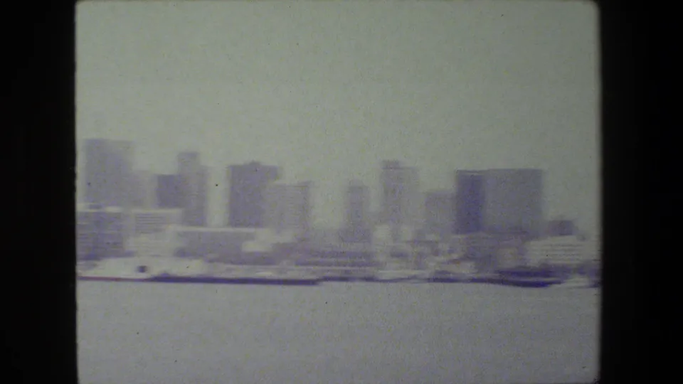 A faded 8mm frame shows the harbor foreground with clustered modern buildings and low waterfront structures in the distance.