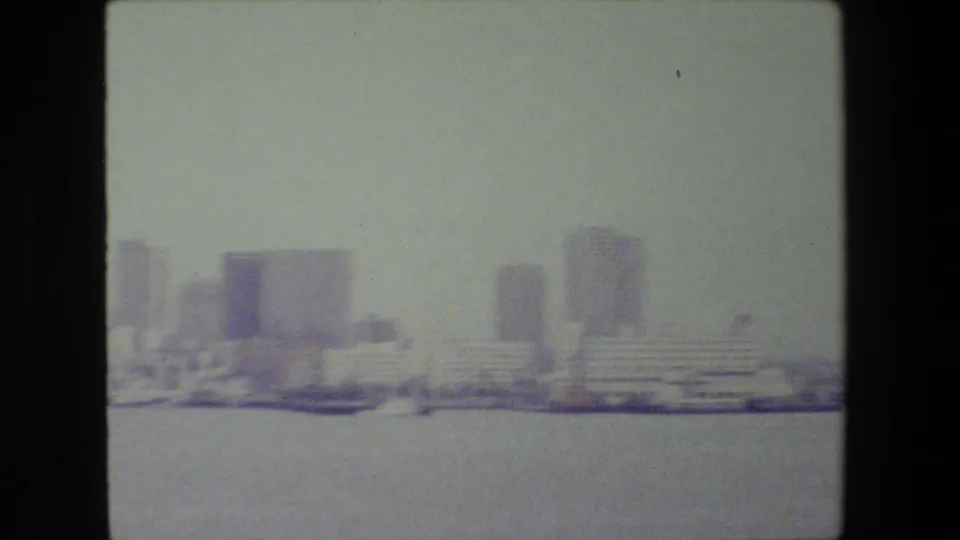 The pan settles on a broad waterfront skyline, including high-rise blocks and a stepped low building beside the water in faded home-movie color.