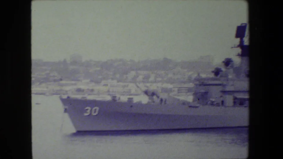 A gray military ship marked with hull number 30 sits in harbor water, with radar equipment and a hazy shoreline visible in the background.