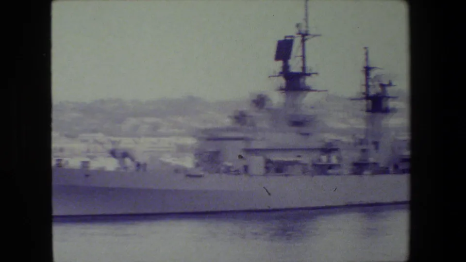 The camera view holds on the side of a gray military vessel, showing deck structures, radar equipment, masts, and a distant hazy city shoreline.