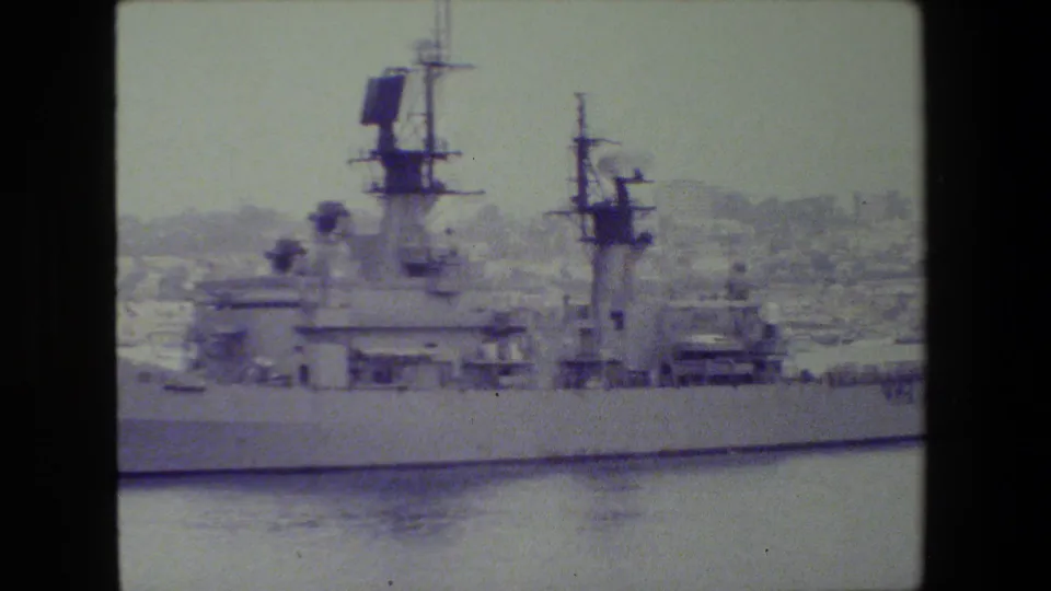 A closer portion of the military ship shows the superstructure, radar panels, mast details, and railings against a hazy harbor backdrop.