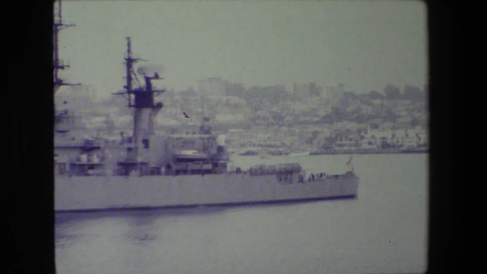The pan reaches the forward section of the gray military ship, with railings, mast elements, calm water, and a hazy shoreline beyond.