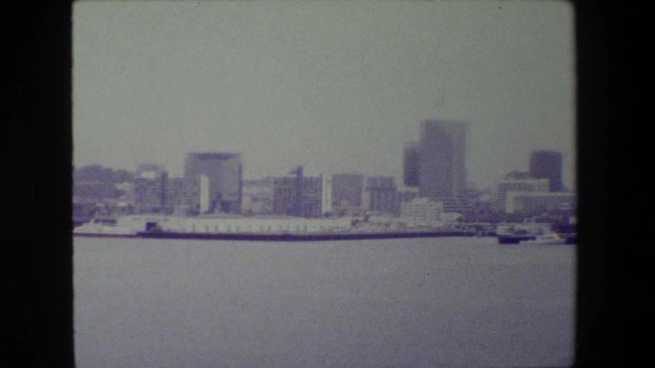 Wide 1981 San Diego harbor view with open water in the foreground, docked ships along the waterfront, and a soft distant skyline of mid-rise and high-rise buildings.