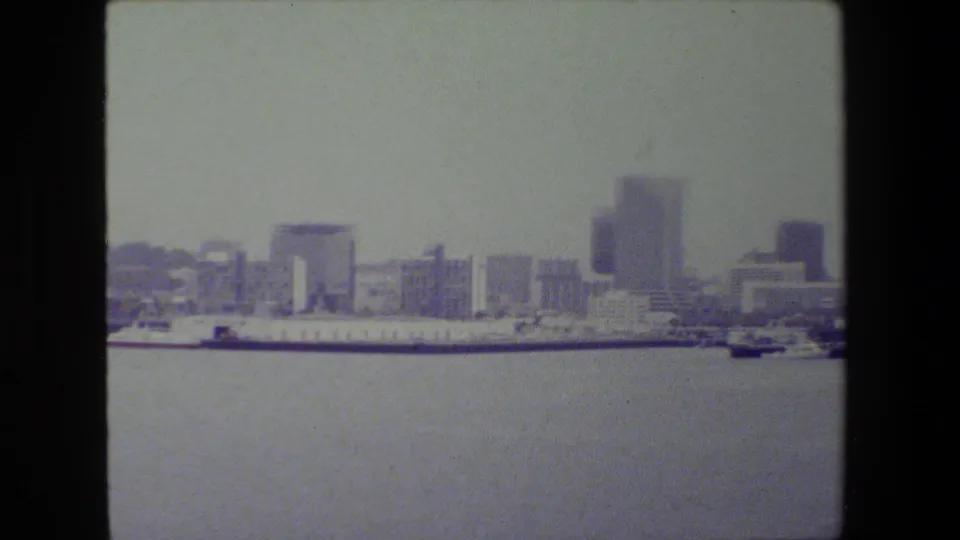 Wide 1981 San Diego harbor view with open water in the foreground, docked ships along the waterfront, and a soft distant skyline of mid-rise and high-rise buildings.