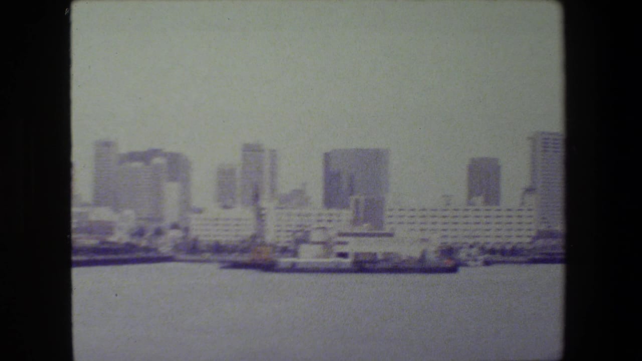 Soft 8mm frame showing a coastal city skyline across harbor water, with a long pale waterfront building, high-rises, and vessels along the shoreline; San Diego is suggested by metadata but not confirmed visually.