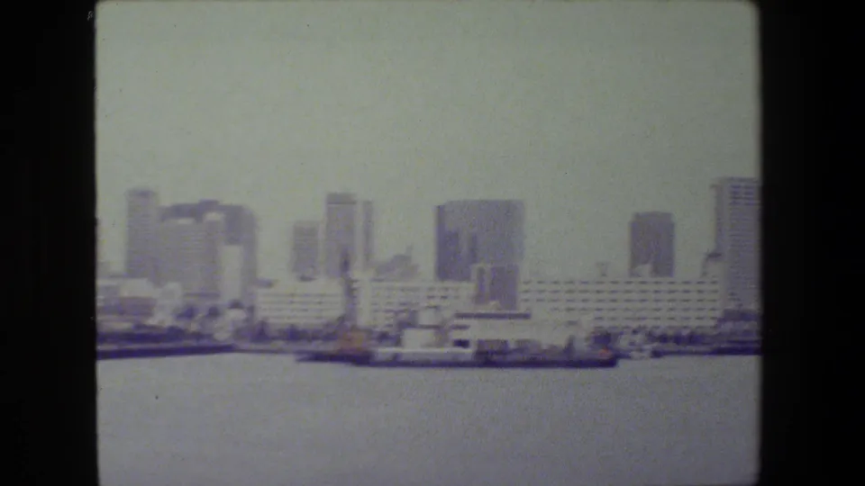 Soft 8mm frame showing a coastal city skyline across harbor water, with a long pale waterfront building, high-rises, and vessels along the shoreline; San Diego is suggested by metadata but not confirmed visually.