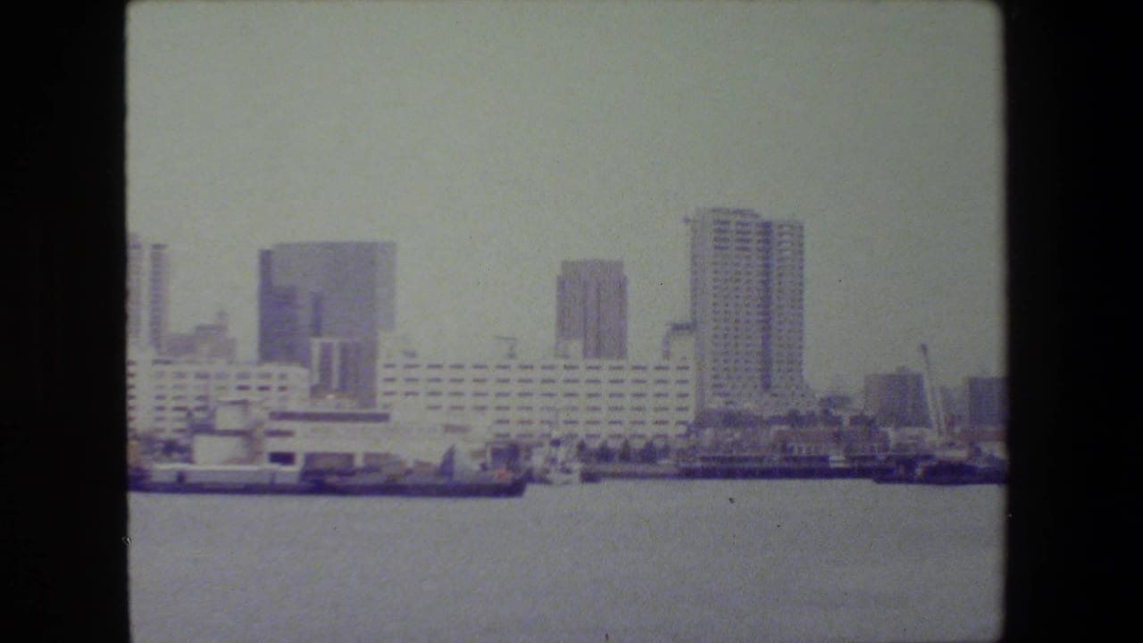 Waterfront city view with a long white building, towers, and boats near the shore.