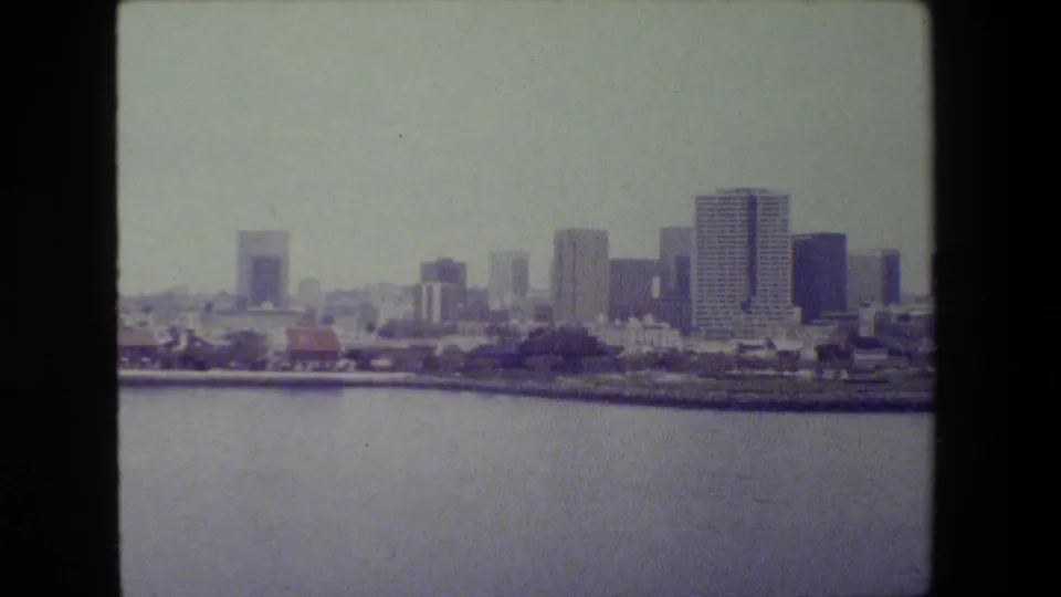 Wider 8mm view across harbor water toward a dense waterfront skyline, including mid-rise structures, taller towers, shoreline development, and a hazy sky.