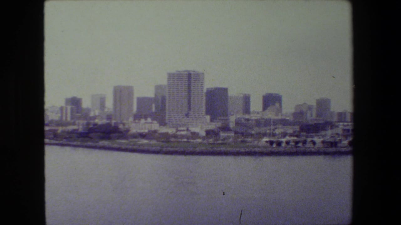 Soft 8mm frame of a distant urban waterfront skyline across calm harbor water, with high-rise buildings and shoreline structures under hazy daylight.