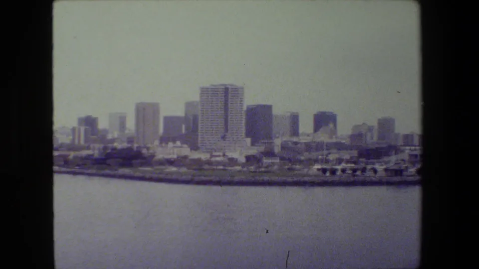 Soft 8mm frame of a distant urban waterfront skyline across calm harbor water, with high-rise buildings and shoreline structures under hazy daylight.