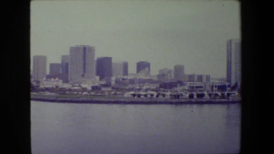 Distant city skyline viewed over harbor water, with multiple high-rise buildings and low waterfront structures in a soft, grainy 8mm frame.
