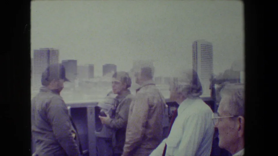 Adults aboard a boat look toward a hazy waterfront skyline, including a man in a military-style cap and others near the rail in soft 8mm color.