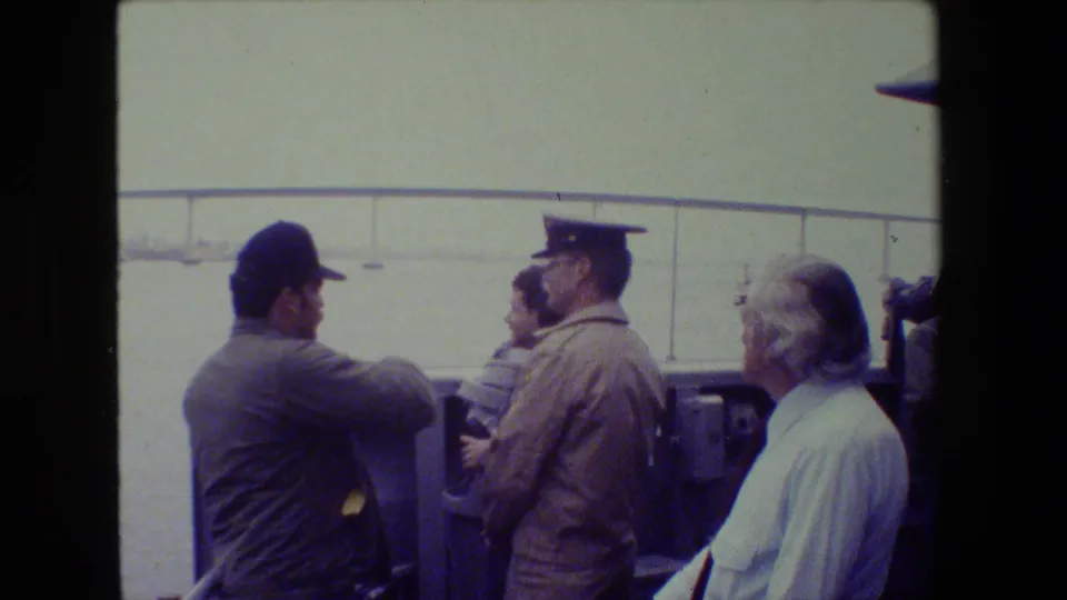 Men aboard a boat stand near the rail as one gestures toward the harbor, with a long bridge and open water visible in the background.